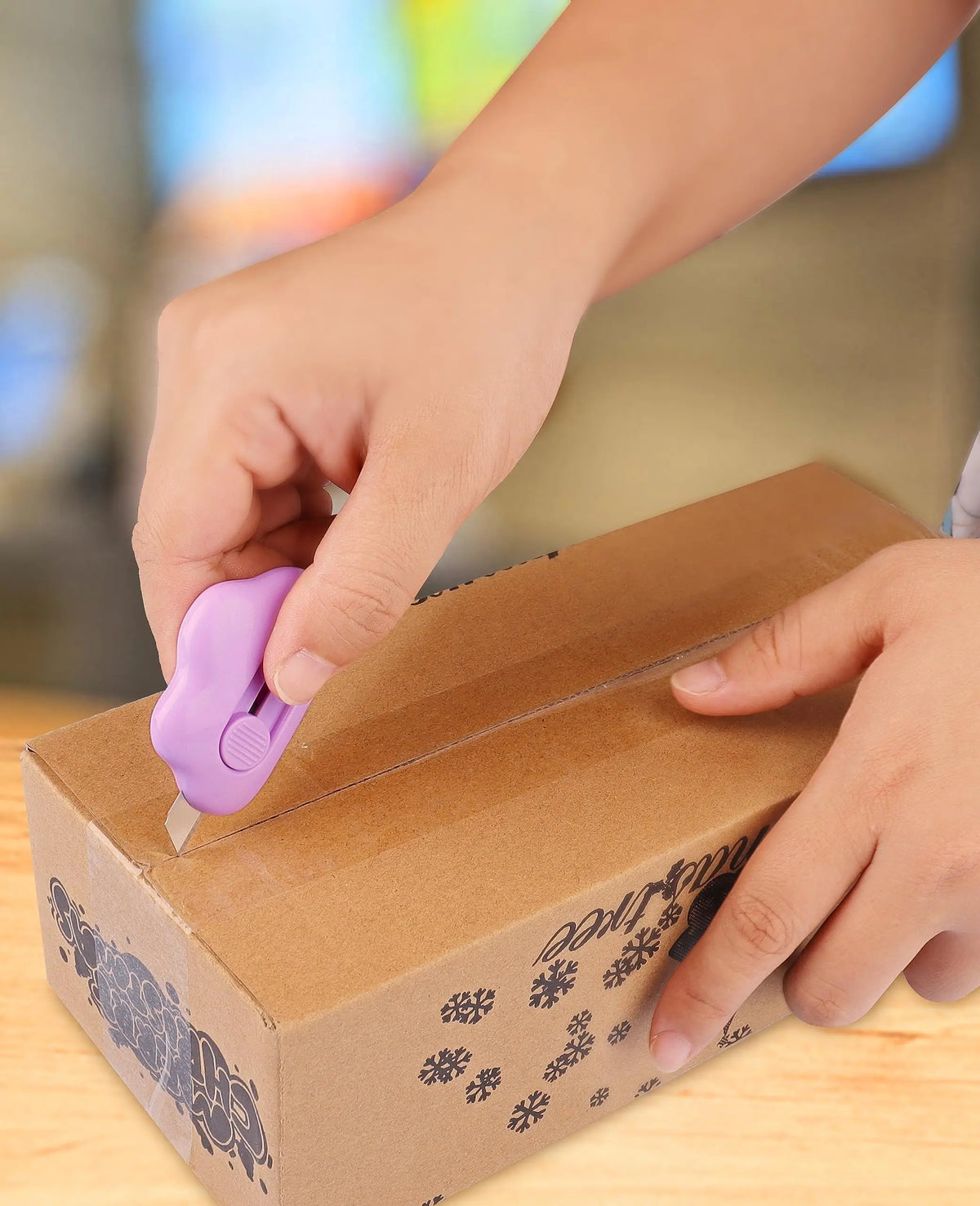 A hand is using a purple cloud-shaped retractable letter opener with a keychain loop to open a cardboard box.