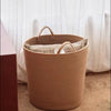 A light tan handwoven cotton rope storage basket with handles sits on a wooden surface.