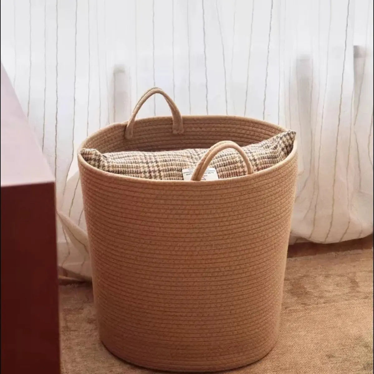 A light tan handwoven cotton rope storage basket with handles sits on a wooden surface.