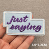 A hand holds a light blue embroidered patch with the text Just saying in purple script.