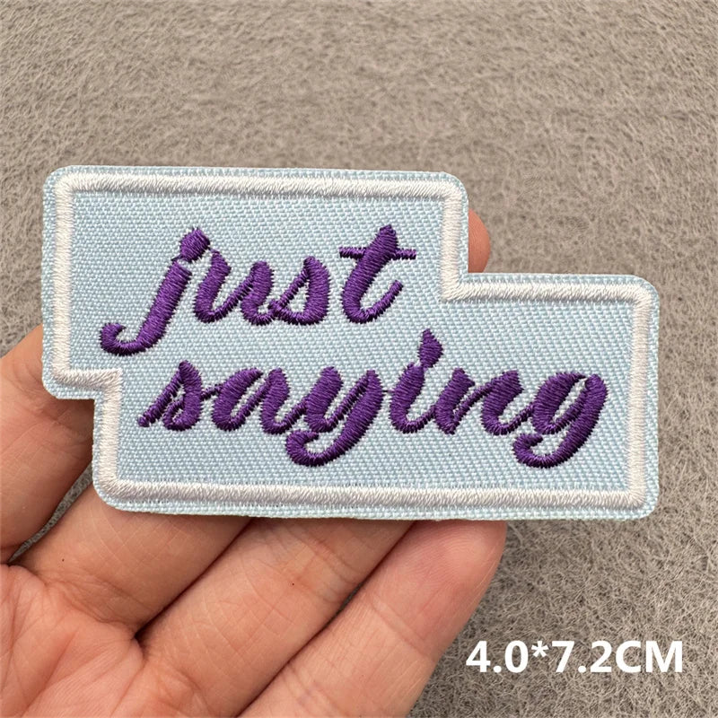 A hand holds a light blue embroidered patch with the text Just saying in purple script.