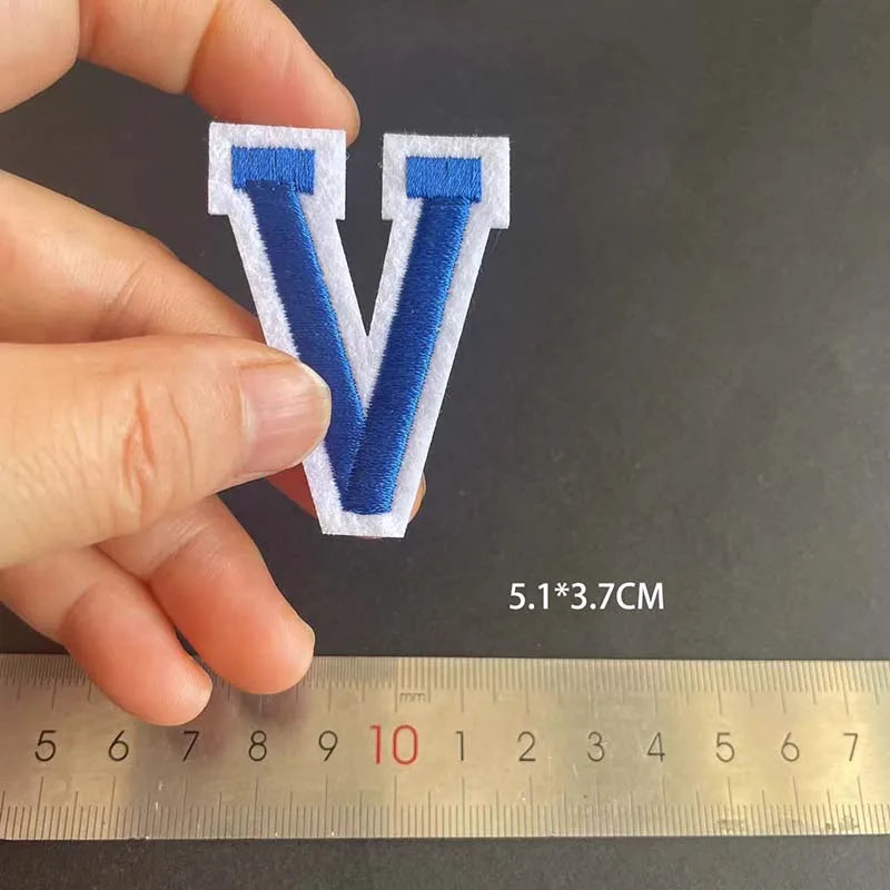 A hand holds a blue embroidered letter V patch on a ruler for scale.
