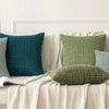 A set of seven textured throw pillows in various shades of green and teal are displayed on a light-colored sofa.
