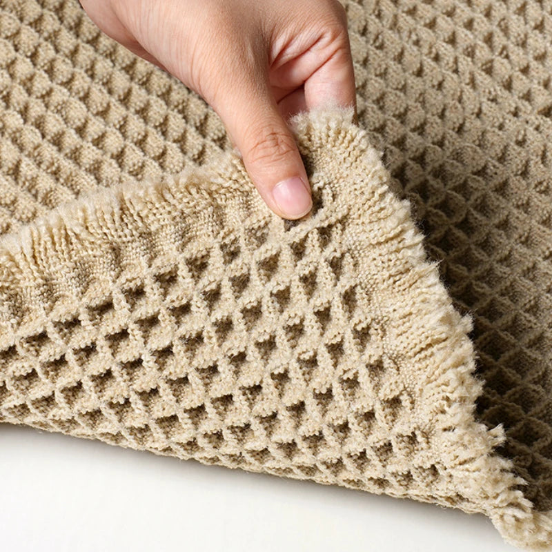A close-up of a beige waffle weave bath mat with fluffy tassels being held by a hand.