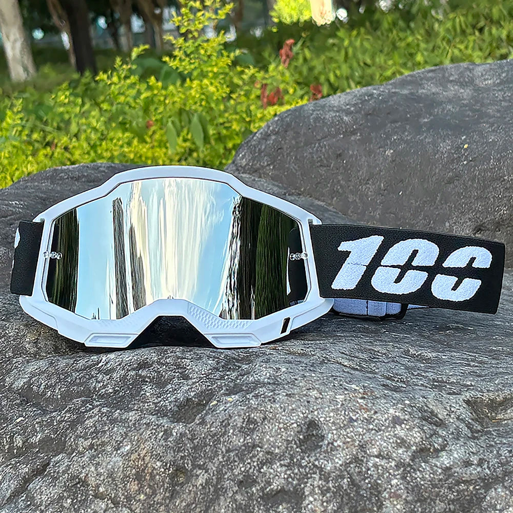 White motocross goggles with a reflective lens and black strap resting on a dark rock.