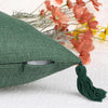 A dark green cotton linen throw pillow cover with a tassel, resting on a white surface next to dried flowers.