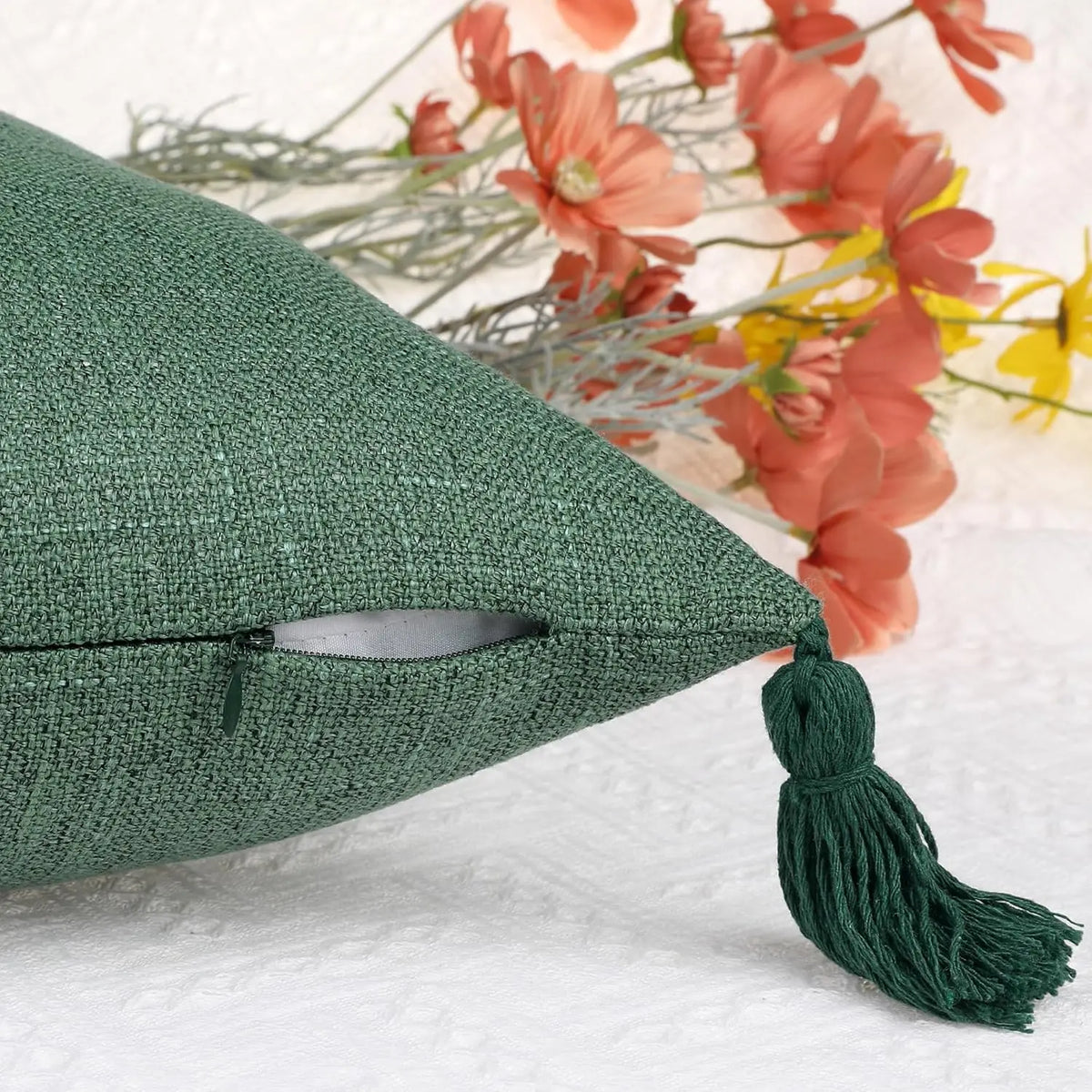 A dark green cotton linen throw pillow cover with a tassel, resting on a white surface next to dried flowers.