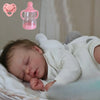 A newborn baby doll sleeping peacefully on a white cloth, with a pink baby bottle and pacifier nearby.