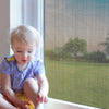 A toddler sits in front of a window with a mesh screen visible, looking out.