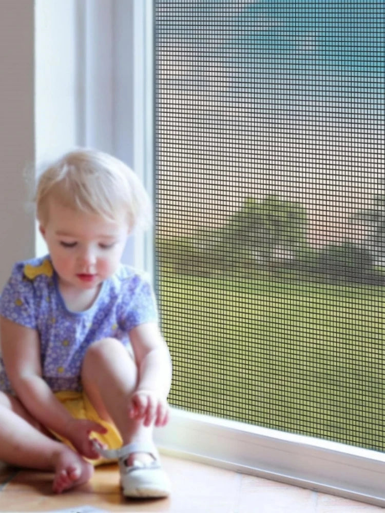 A toddler sits in front of a window with a mesh screen visible, looking out.
