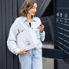 A woman in a denim shirt holds a white poly mailer box and looks at her smartphone.