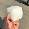 A hand holds a white, square squishy stress relief toy made of milk tofu against a gray concrete background.