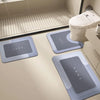 Three gray diatomium mud floor mats placed on a light beige bathroom floor near a toilet.