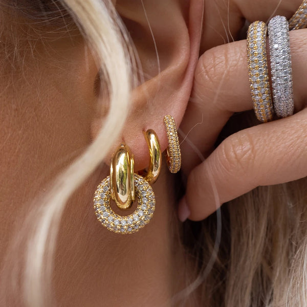 Gros plan d'une main portant des clous d'oreilles en forme de boucle dorée géométrique, contre une peau claire et des cheveux