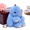 A fluffy blue angel bunny keychain with a pom pom tail resting on a dark box next to a pink rose.