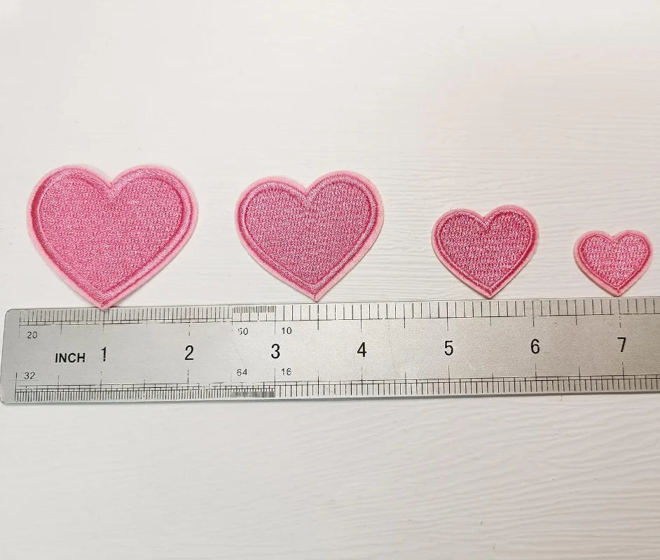 A row of five pink embroidered heart iron-on patches laid out next to a ruler.
