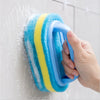 A hand is holding a blue and yellow sponge cleaning brush against a white wall.