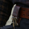 A close-up of a brown leather tool holster hook with a steel J-hook attached to a dark fabric belt.