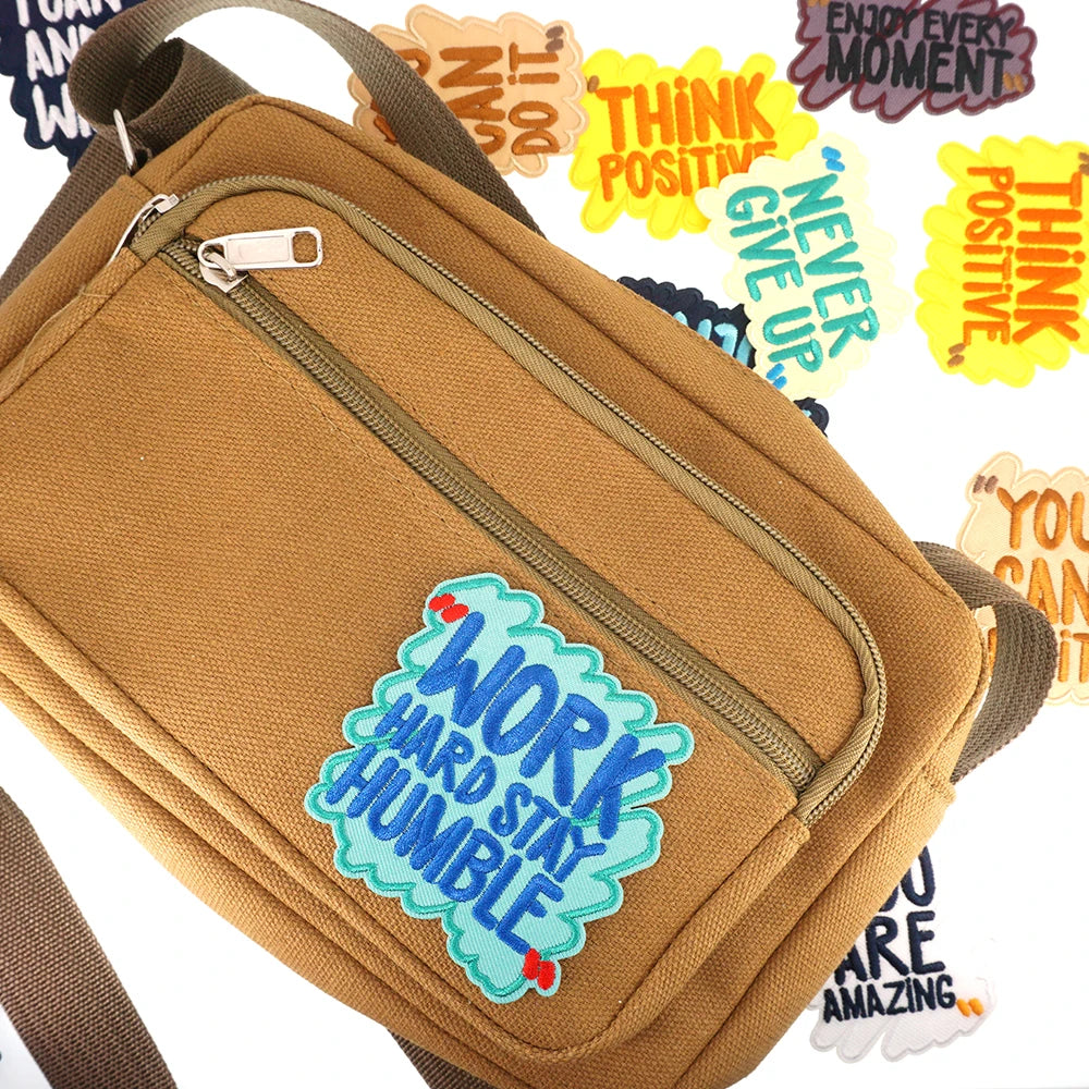 A tan canvas bag with a zippered pocket featuring embroidered humorous iron-on patches encouraging positivity and hard work.