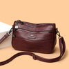 A maroon leather compact square crossbody bag with a zipper and leather strap resting on a light background.