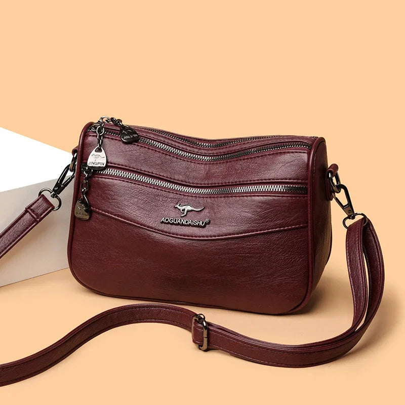A maroon leather compact square crossbody bag with a zipper and leather strap resting on a light background.