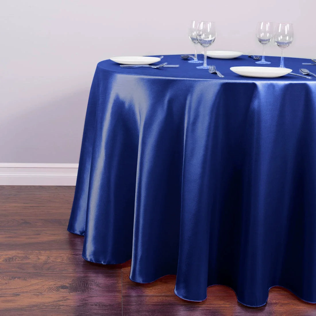 A deep blue satin tablecloth is set for an event with white plates and glassware on a wooden table.