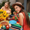 A woman laughing while eating a tomato in an orange shirt next to a colorful Mexican serape table runner.