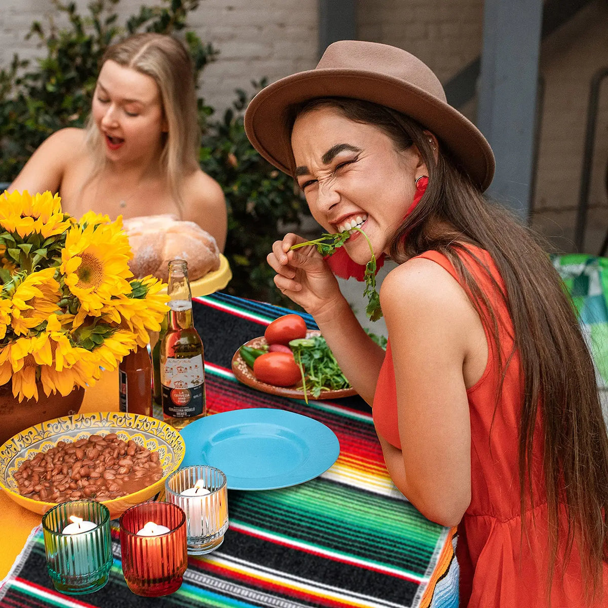 A woman laughing while eating a tomato in an orange shirt next to a colorful Mexican serape table runner.