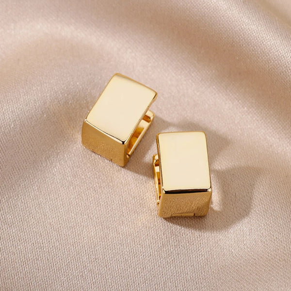 Un par de pendientes de aro geométricos cuadrados en metal dorado pulido sobre un fondo de tela beige.