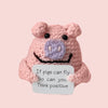 A handmade crochet doll shaped like a pink pig with purple features and a black mouth, featuring a positive message.
