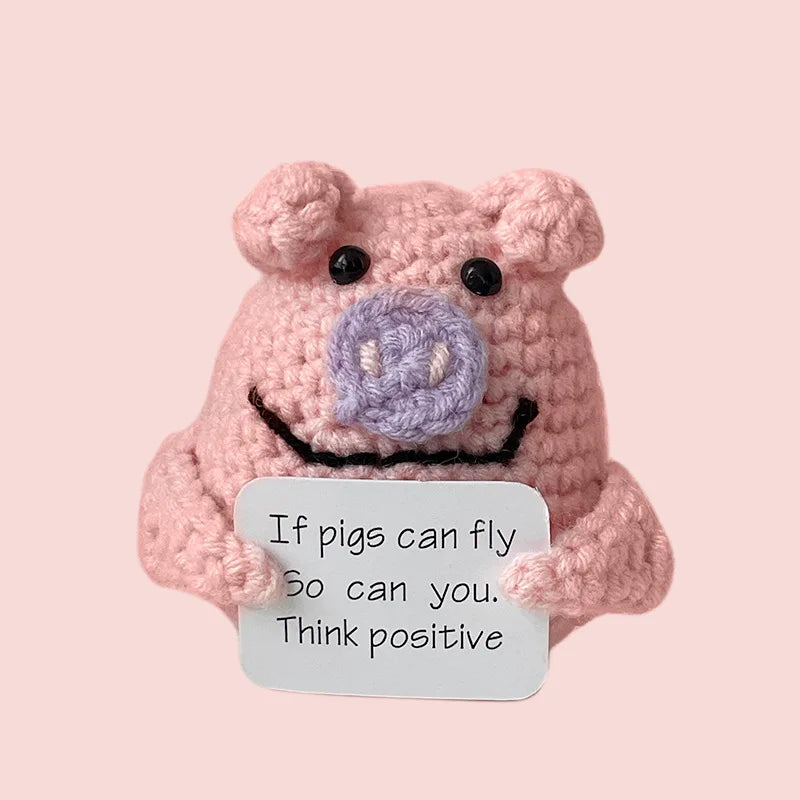 A handmade crochet doll shaped like a pink pig with purple features and a black mouth, featuring a positive message.