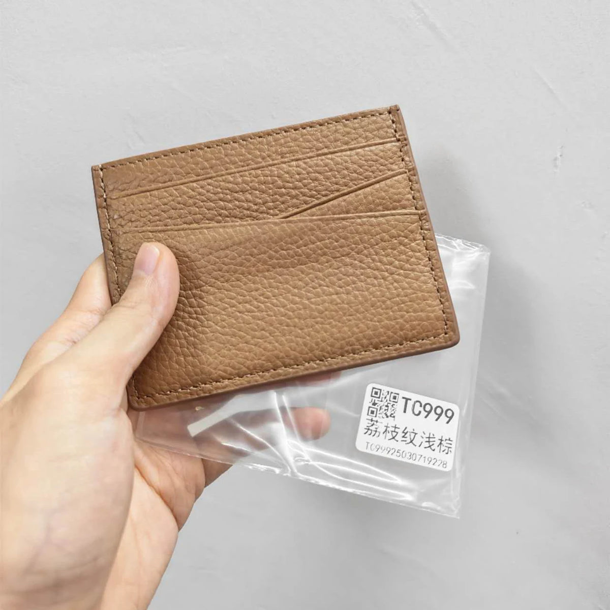 A hand holds a brown genuine leather slim card holder wallet packaged in clear plastic.