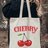 A white canvas tote bag with red cherry print and text on it, held by a person outdoors.