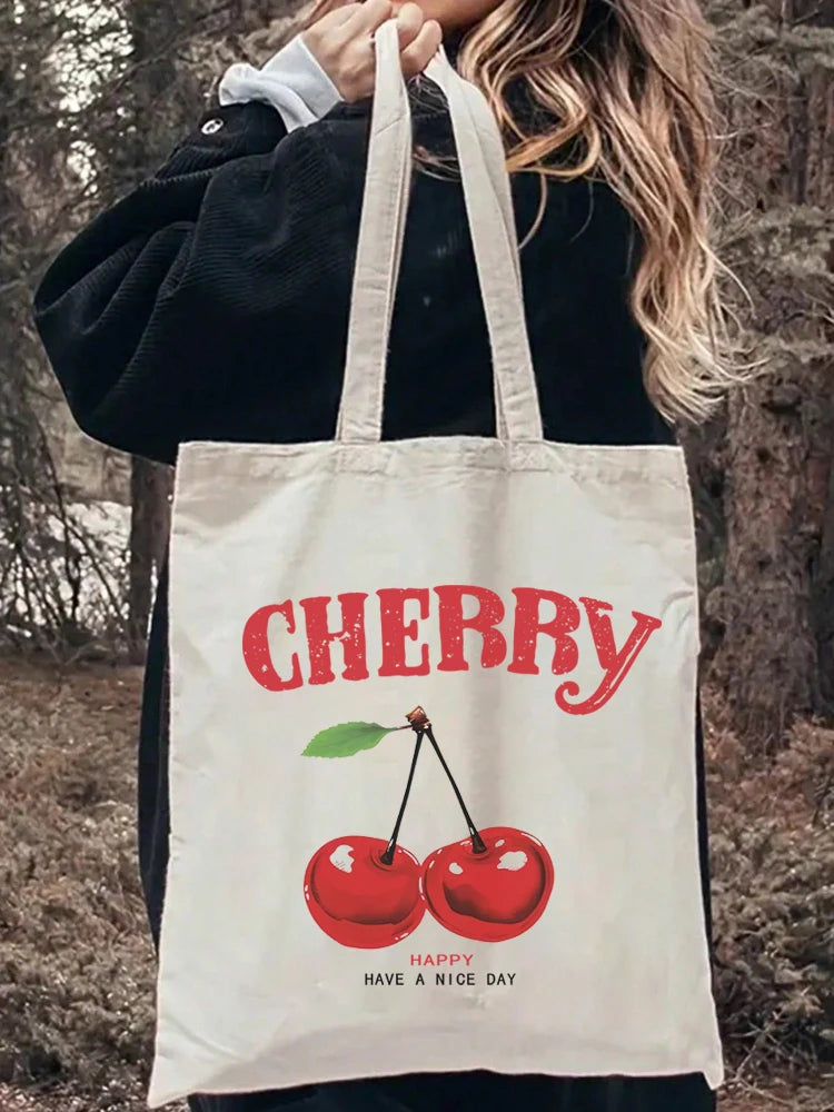 A white canvas tote bag with red cherry print and text on it, held by a person outdoors.