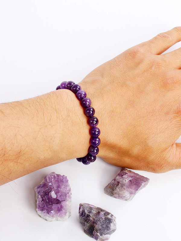 Una mano vistiendo una pulsera de cristal amatista púrpura junto a varias piedras de amatista crudas.