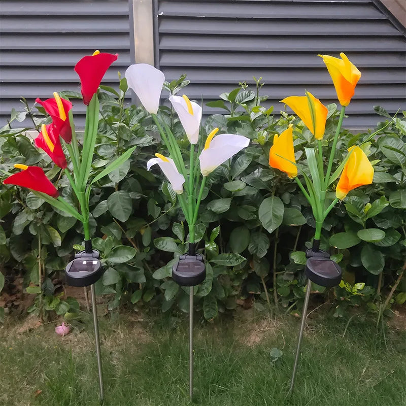 A row of potted calla lilies with red and yellow blooms illuminated by solar-powered LED lights against a green hedge.