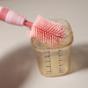 A silicone baby bottle cleaning set with a pink brush is shown dipping into a clear measuring cup.