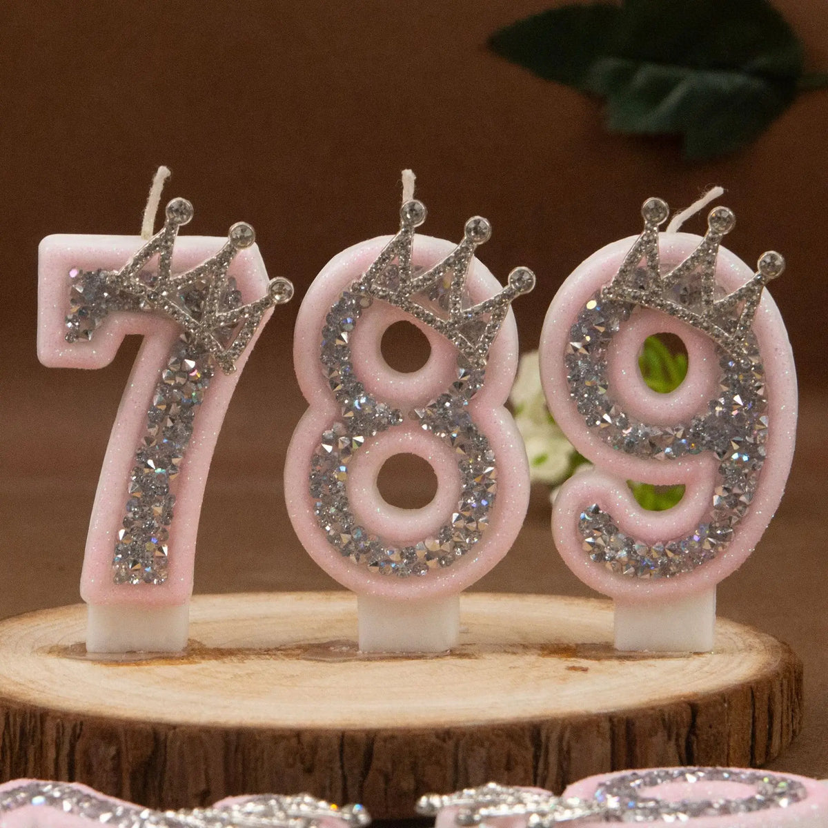 Three pink rhinestone crown number candles for birthday cakes, numbered 789, displayed on a wooden surface.