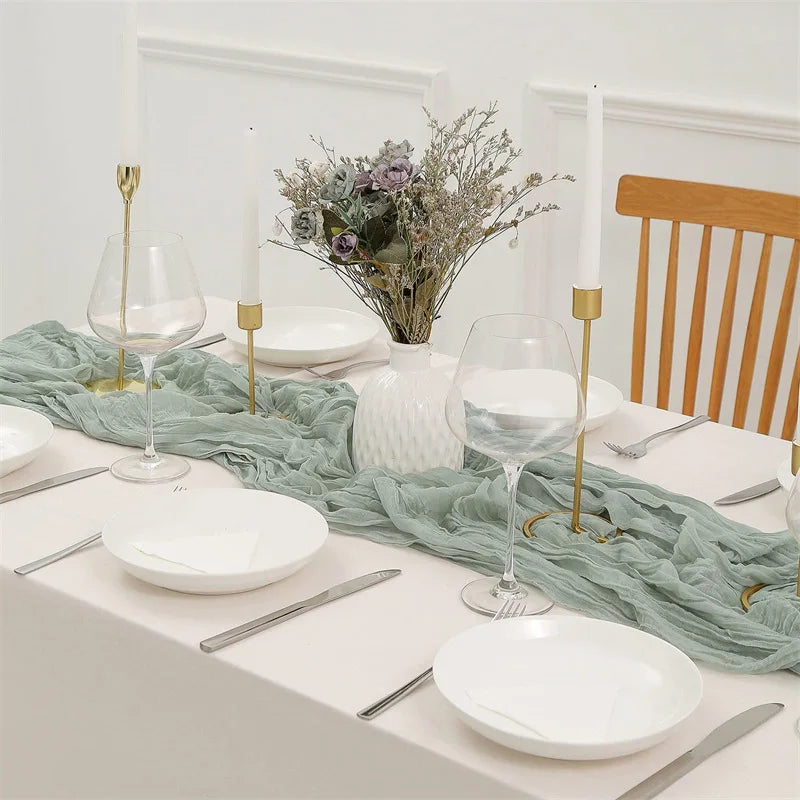 A white table setting featuring a sage green cotton gauze table runner, white plates, silverware, and tall candles with dried