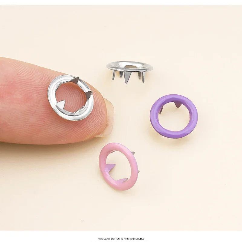 Un juego de cinco pinzas de botón en uñas de plata, púrpura y rosa mostrados en un dedo.