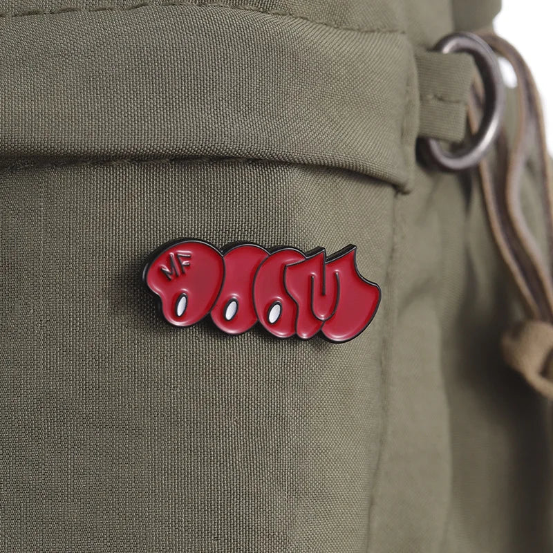 A red bubble enamel pin brooch with the text MF DOOM is attached to a military green fabric.