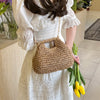 A woman wearing a white dress holds a woven straw chain crossbody handbag and green stalks of flowers.