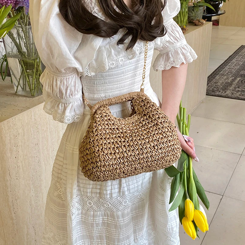 A woman wearing a white dress holds a woven straw chain crossbody handbag and green stalks of flowers.