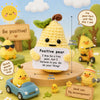 A yellow crochet pocket hug doll sits on a wooden surface surrounded by small plants and encouraging signs.