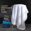 A white luxury bath towel draped over a stand, featuring gold embroidered stripes.