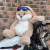 A fluffy, light brown rabbit plush toy with white accents and sunglasses is resting on a black motorcycle.