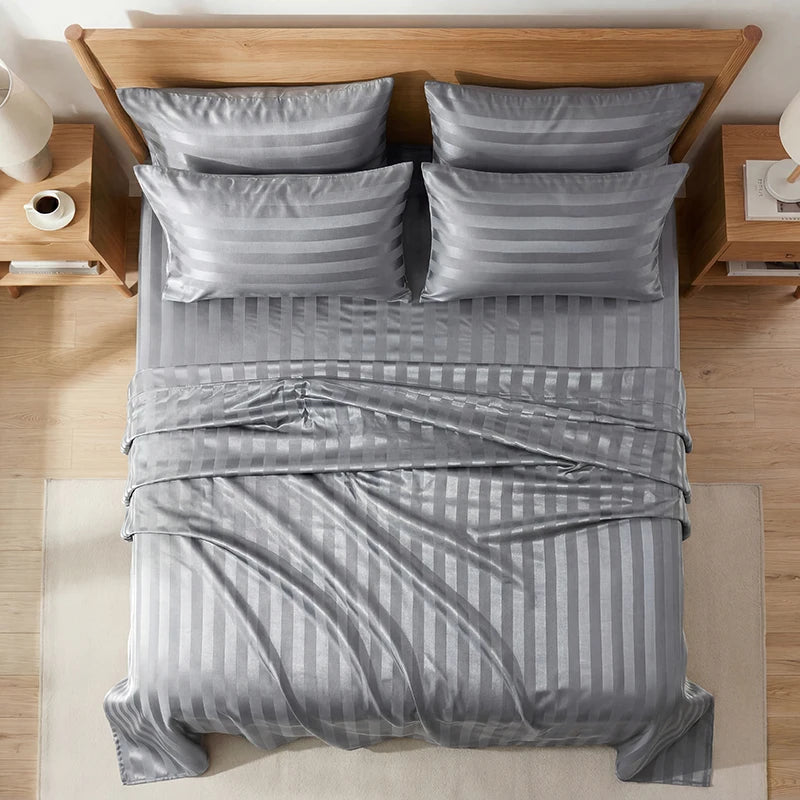 A set of four striped satin bed sheets and pillows in gray are displayed on a wooden bed frame.