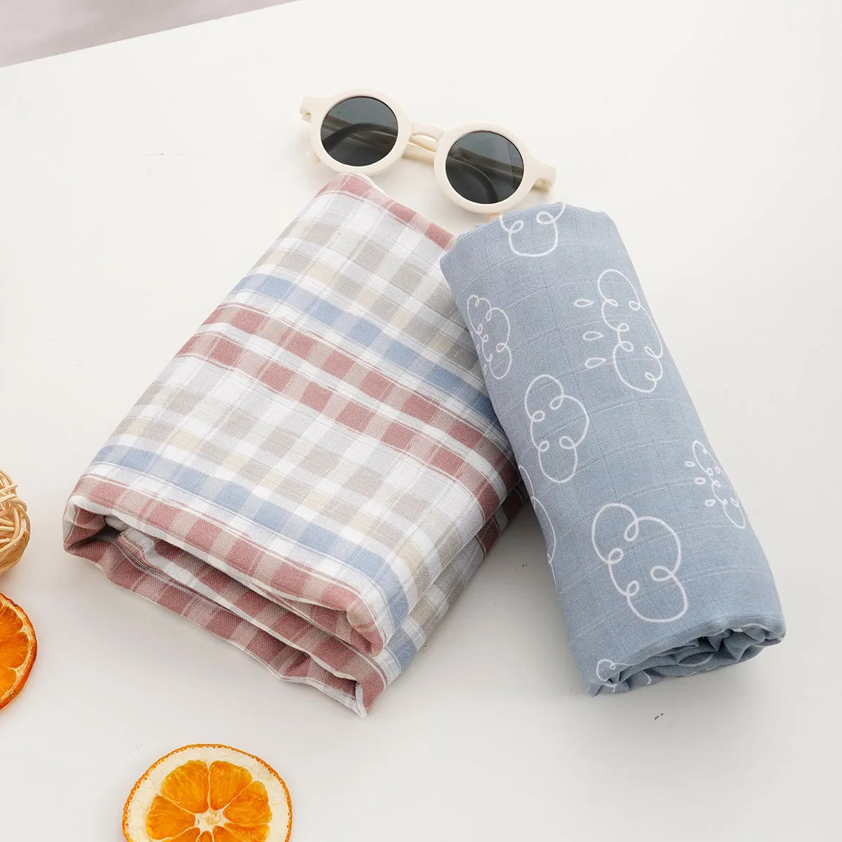A light blue muslin baby swaddle blanket rolled up next to a pair of sunglasses and an orange slice.