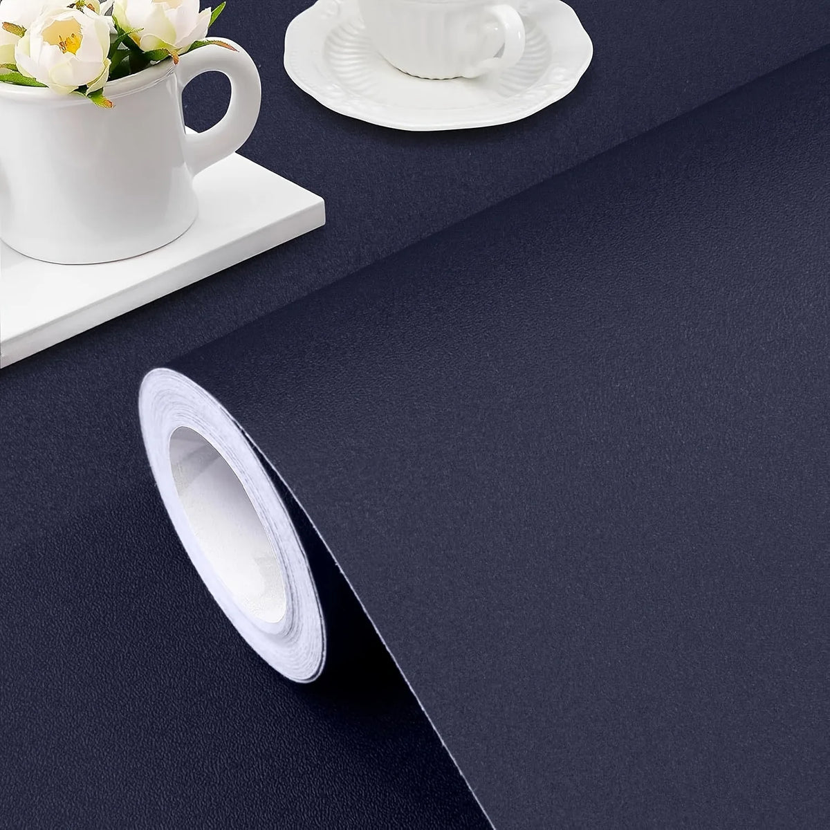 A roll of white self-adhesive peel-and-stick wallpaper lying on a dark blue surface next to a white cup and saucer.