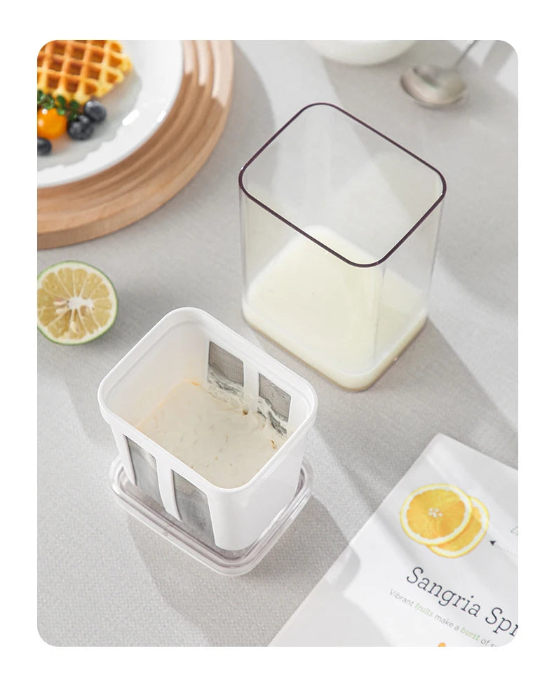 A clear stainless steel yogurt strainer with a fine mesh filter is shown next to a glass of milk and an orange slice.